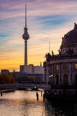 Sunset view of the Berlin TV Tower (Fernsehturm) rising behind the domed Bode Museum, with a bridge over the Spree river, colorful sky, and a jet contrail crossing above the city. © That French Bloke