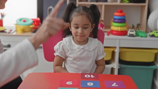 Little girl student at a table with number cards as woman teacher points finger to cards in a playroom building; concentration.