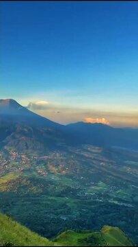 Bright blue sky stretches clear and wide, twin volcanic peaks rise above hazy valleys, green hills and small town spread far below