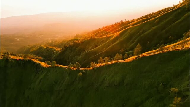 Sunlit rolling green hills glow warm gold, scattered shrubs and trees dot slopes, soft white clouds drift far above misty distant valleys