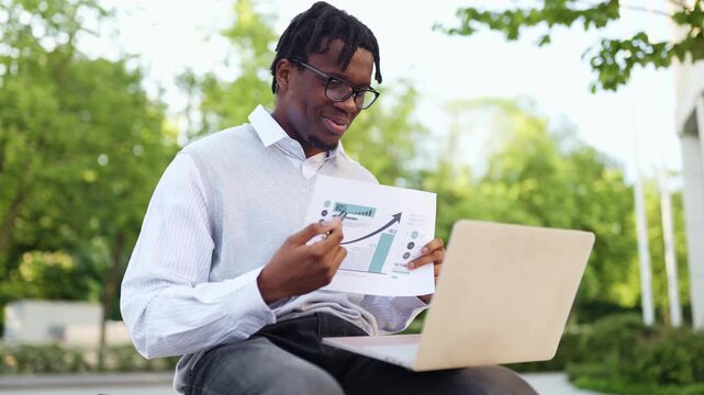 A smiling african american man in glasses reviews a financial report with a laptop in a park setting