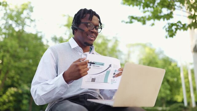 A focused african american man wearing glasses and a headset explains a bar graph with an upward trend to a laptop screen in a park setting