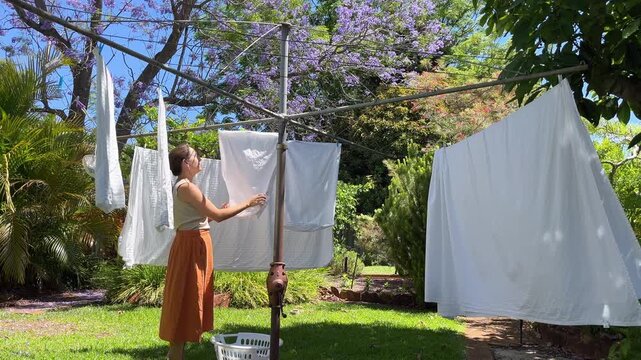 Woman hanging fresh laundry to dry on a rotating clothesline in a green backyard garden under a bright blue spring sky