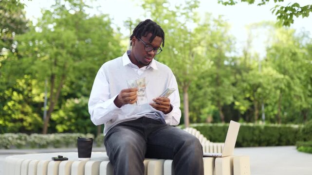 A young african american man wearing glasses sits on a bench in a park and counts a stack of hundred dollar bills while a laptop is nearby