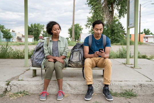 Young budget travelers with backpacks waiting at bus stop, using smartphone for public transport while backpacking on an affordable journey