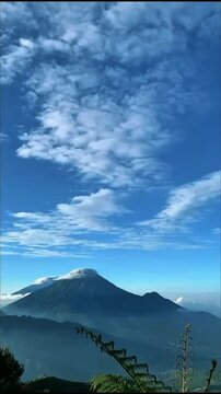 Twin volcanic peaks capped with thin clouds stand above layered misty ranges, bright blue sky filled with fluffy drifting white clouds