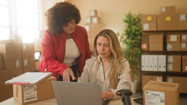 Woman points at laptop amid stacked parcels and clipboard while colleague sits in a packing room building; collaboration joy.
