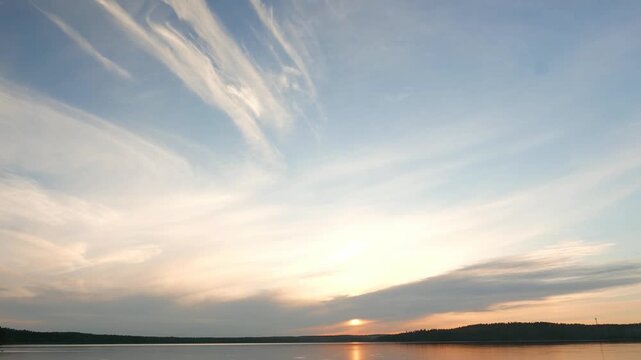 Light stretched clouds over evening sky, lake sunset skies time lapse shot. Sun disk ahead, hide behind dense stripe of clouds, move down to horizon line. Reflex on flat water. Fly quickly above,