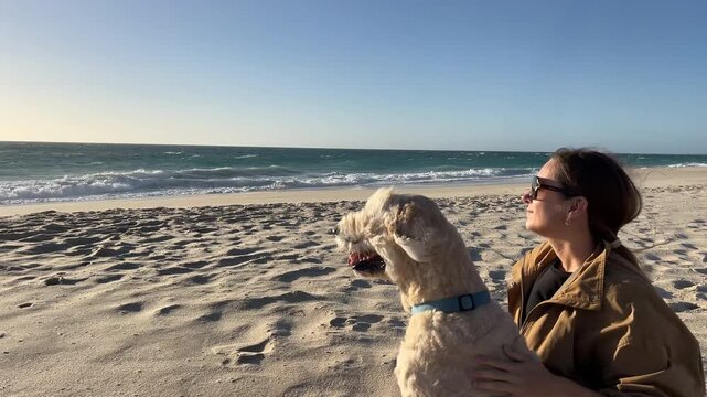 Happy woman wearing sunglasses pets her fluffy dog while relaxing on the sandy beach during a sunny afternoon by the ocean.