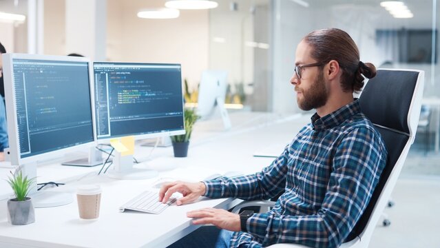 Close up of happy young Caucasian IT specialist sitting at desk in office coding while typing on computer and smiling at camera. Male programmer in cabinet looking at camera. Internet technology job