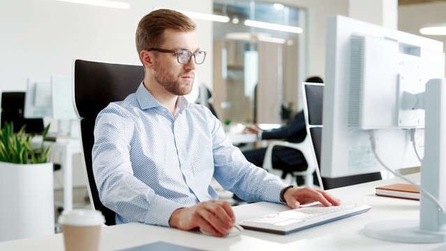 Handsome concentrated businessman sitting in modern cabinet and looking at monitor typing on computer keyboard at workplace. Close up of young Caucasian man in glasses working on computer