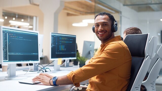 Camera approaching young Hindu man IT programer working on desktop computers typing on keyboard coding and listening to music in headphones. Close up of professional worker in modern office