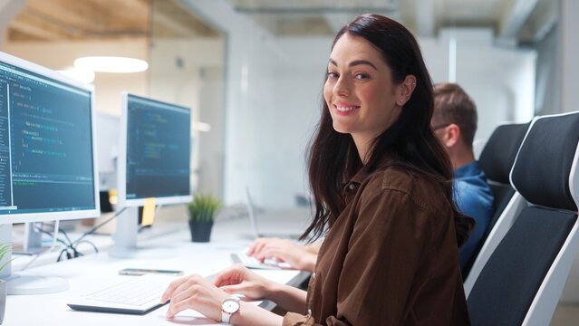 Rear of young pretty woman IT programer working on desktop computers typing on keyboard coding a program turning to camera and smiling. Close up of professional worker in modern tech company