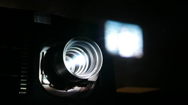 Motion picture projector lens, power beam shine through glass and optic. Blurred image projected on booth window, extreme close up view of kino beamer at work