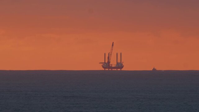 Industrial oil platform operating at sea while a small support vessel sails nearby during a colorful evening sunset