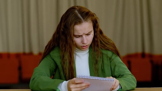 Teenage girl in green blazer reading notebook with finger line by line, cramming text before exam and feeling stressed about difficult school test.