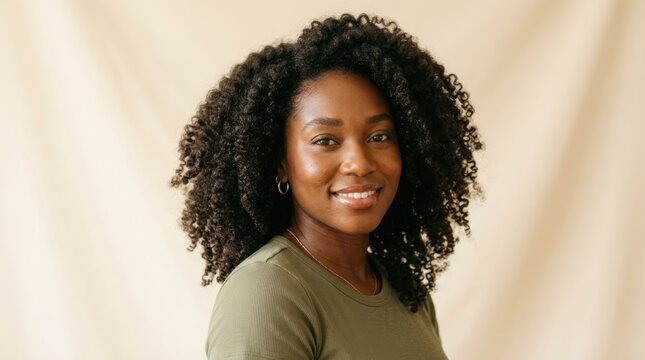 A woman with curly hair smiling, wearing a green shirt, standing in front of a beige background.