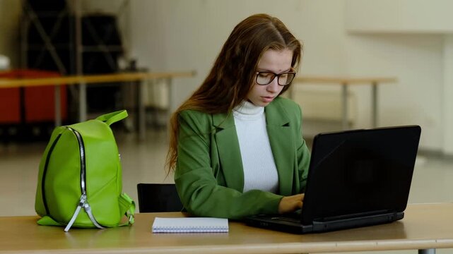 Teen girl with long hair and stylish green suit typing on laptop at school desk, notebook and backpack nearby, concept modern education and digital learning for teenagers.
