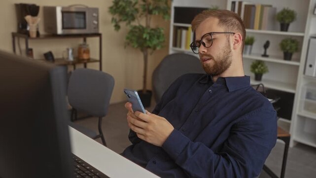 Man with glasses and beard holding smartphone with both hands while leaning back in office chair near desktop monitor; boredom distraction.