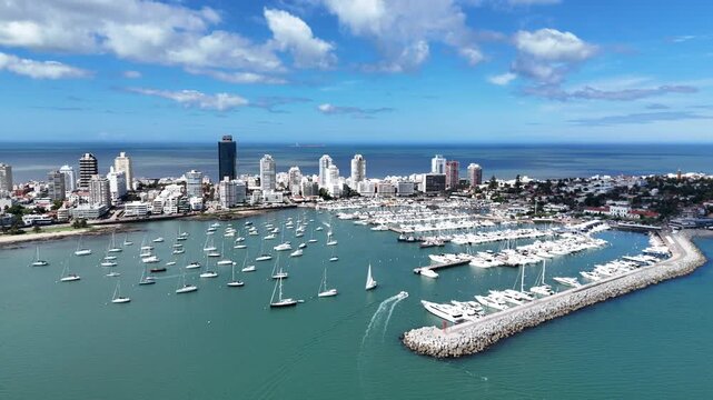 Aerial drone footage of sailboats navigating Punta del Este Marina with luxury yachts and the city skyline in the background.