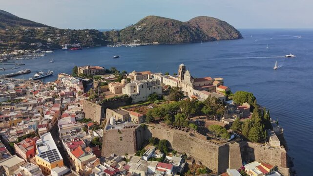 Aerial view of ancient Lipari castle fortress sitting on high coastal cliff. Colorful Mediterranean town houses stretch around historic port. Calm blue waters of Aeolian islands, Lipari