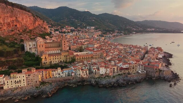 Drone shot of historic Cefalu town bathed in warm sunset golden hour light. The iconic cathedral and La Rocca cliff rise above coastal houses. Cefalu, Sicily, Italy at sunset