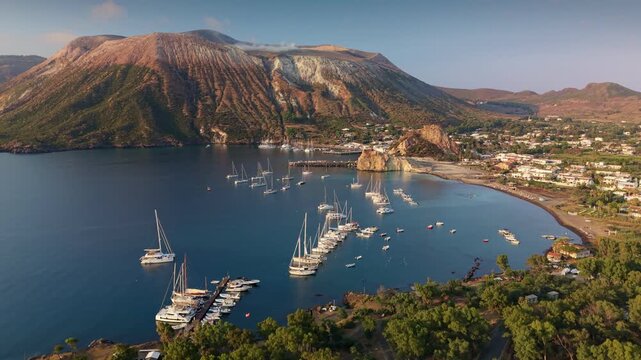 Aerial view of massive Gran Cratere volcano on Vulcano island during golden hour sunset. Sailboats lie at anchor in calm bay at mountain foot. Volcanic peak in Sicily, Aeolian archipelago, Italy