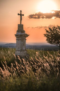 An old stone wayside cross standing in a tall grass field during a dramatic orange sunset in the countryside.