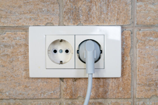 White electrical wall socket with a plugged in power cord mounted on a beige tiled wall in a modern home interior setup