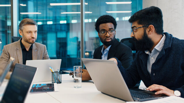 Three male professionals collaborating during business meeting at office table, one explaining data on laptop. Team focused on digital strategy, charts and tech devices visible around.