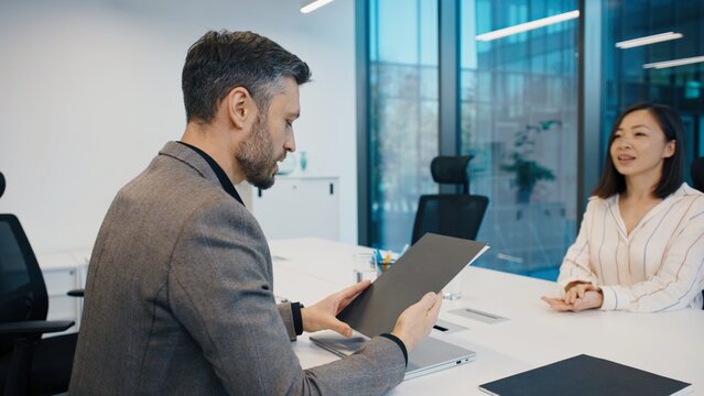 Man in blazer reading documents during meeting with Asian woman in striped blouse at white desk. Professional office environment with glass walls and modern furniture. New employee interview. CV.