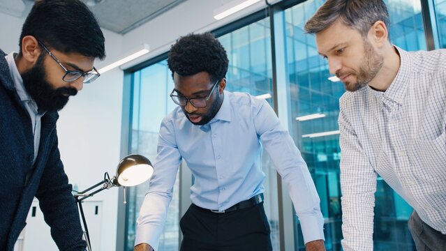 African American male explaining concept to South Asian and Caucasian colleagues during discussion in office. Three professional men planning wind energy model project near window. Indoors.