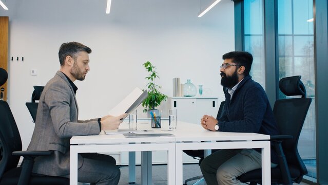 Caucasian man in suit shaking hands with South Asian man during job interview. Confident gesture at meeting in modern office. CV or resume reading. Interviewing for new work position in company.