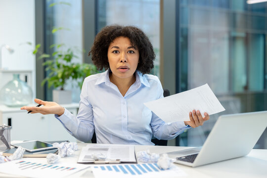 Portrait of a young African American woman sitting at an office desk, looking worriedly at the camera, holding documents and spreading her arms in frustration