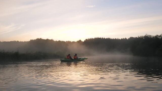 Two people in kayak paddling on calm river surrounded by morning mist. Sunrise creating a serene and picturesque landscape with a forest silhouette in background. Mystical mist over surface of river