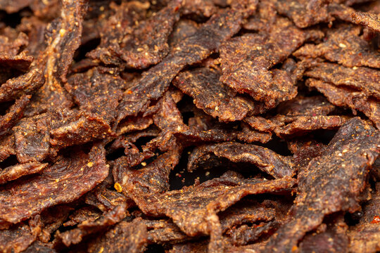 Textured macro shot of seasoned dehydrated meat jerky. Homemade smoked beef jerky strips piled tightly together, creating a rich organic food pattern with visible seasoning flakes.