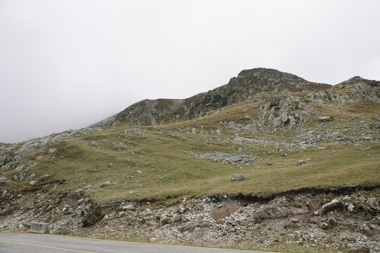 Transalpina mountain pass landscape with rocky peak, alpine vegetation, and road under overcast cloudy sky in Par&acirc;ng Mountains, Romania
