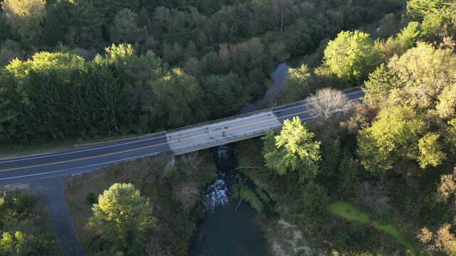 Concrete bridge spans two side of a narrow river valley. A flowing creek rushes of rocks and rapids in a thick green forested natural area in the Midwest, USA. Aerial video
