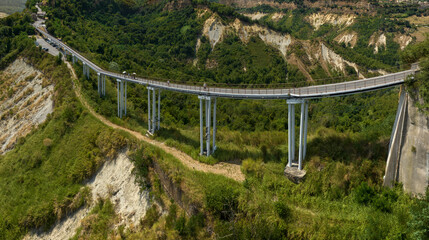 Fototapeta premium Aerial view of the long pedestrian bridge connecting Civita di Bagnoregio, stretching across a steep green valley and clay badlands with eroded slopes in Italy. It's located in Lazio region.
