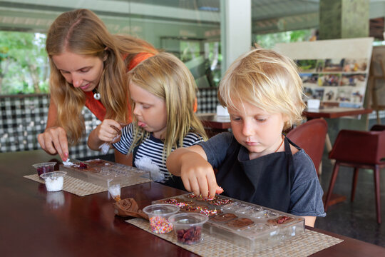 Kids and their mother participate in a chocolate making workshop in Bali, decorating sweets with toppings. A fun and educational family activity during tropical holidays.