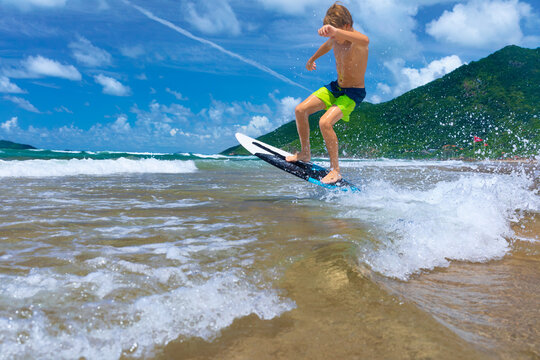 Young boy running, jumping on skimboard and sliding by shallow water on sunny sea beach. Skimboarding, childhood adventure, active lifestyle, watersports, summer vacation and fun outdoor activities.