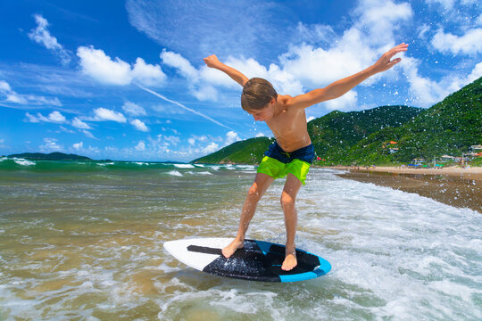 Young boy running, jumping on skimboard and sliding by shallow water on sunny sea beach. Skimboarding, childhood adventure, active lifestyle, watersports, summer vacation and fun outdoor activities.