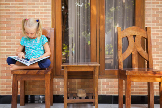 Little girl sitting and reading book at tropical house terrace. Peaceful moment of travel, learning, and childhood discovery under natural light. Ideal for education, lifestyle, family tourism themes.
