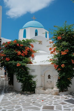 Iconic white Greek church with a blue dome surrounded by blooming flowers, showcasing classic Cycladic architecture beneath a bright Mediterranean sky.