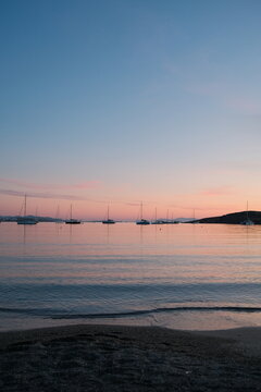 Soft pastel sunset over Paralia Parikia in Paros, Greece, with anchored sailboats, calm water, and gentle waves creating a peaceful Mediterranean seascape.
