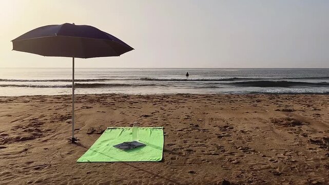 Beach umbrella planted in the golden sand, with a towel underneath holding a book and a glass bottle of fresh water. In the distance, the silhouette of a woman enjoys swimming in the sea