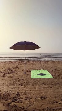 Vertical close up of a beach umbrella planted in golden sand, with a towel underneath holding a book and a glass bottle of fresh water. In the distance, the silhouette of a woman swimming in the sea 