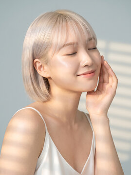 Beautiful asian woman with platinum blonde bob and bangs closing eyes while touching soft radiant skin in bright daylight with striped shadows on face for skincare advertisement shoot.
