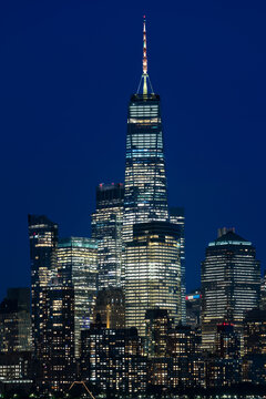 Vertical night view of downtown Manhattan featuring brightly lit high rise skyscrapers against a deep blue evening sky in New York.
