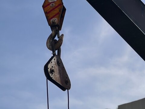 A heavy-duty industrial crane hook with a pulley system suspended against a clear blue sky, showing signs of wear and metallic texture from construction or shipping operations.